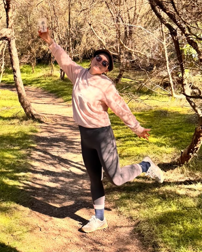 Claire Jingyuan Ye standing on a sunlit bush track with one arm raised, surrounded by trees
